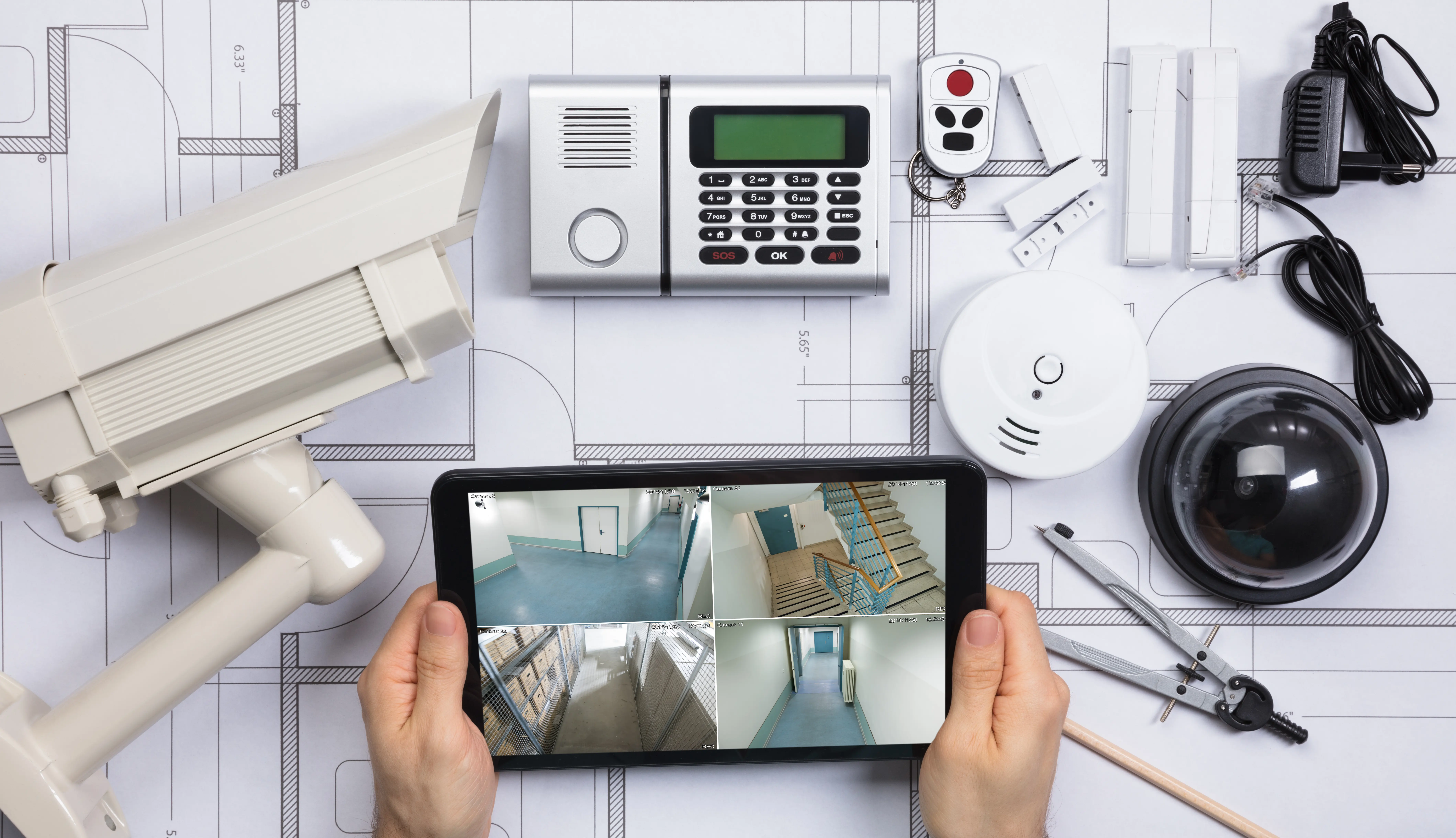 Image shows a security systems like CCTV, Fire Alarm, Carbon Monoxide Alarm, Burglar Alarm and remote locking device on a table with project drawings.