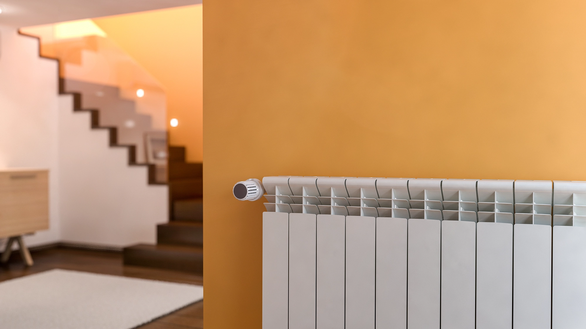 Image of a white radiator with thermostatic valves located in an amber-colored room with a luxurious staircase, highlighting the elegant interior design.